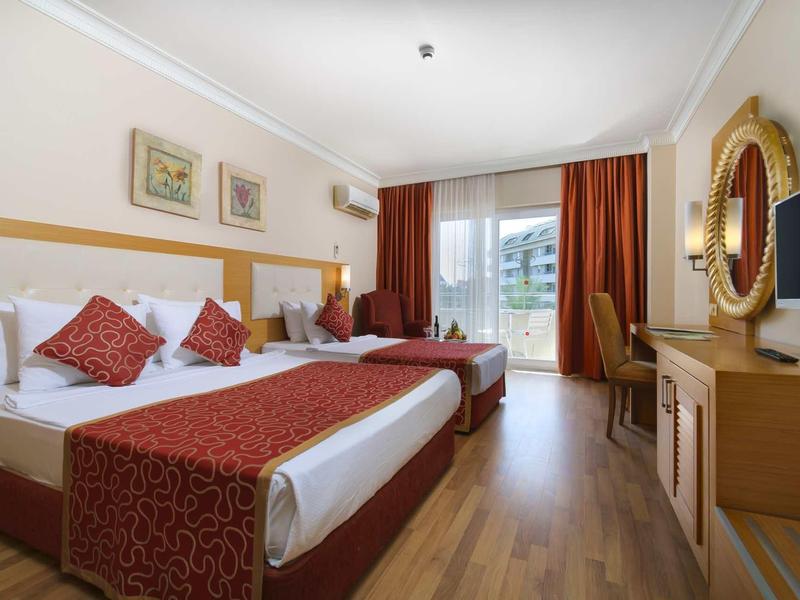 Světle zařízený hotelový pokoj s manželskou postelí, červeným povlečením a velkým zrcadlem.