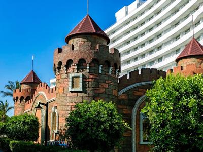 Mauer aus roten Steinen mit runden Türmen vor modernem, weißem Gebäude und blauem Himmel.