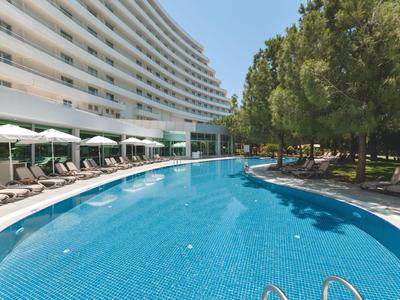 Piscina rotonda dell'hotel con lettini e alberi verdi, edificio moderno a più piani sullo sfondo.