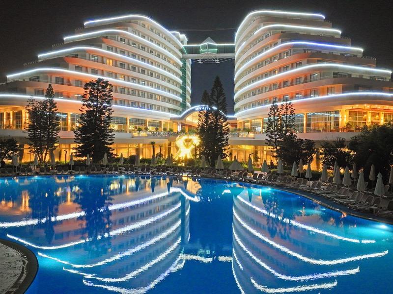 Hotel moderno iluminado con piscina y reflejo por la noche, rodeado de árboles y tumbonas.