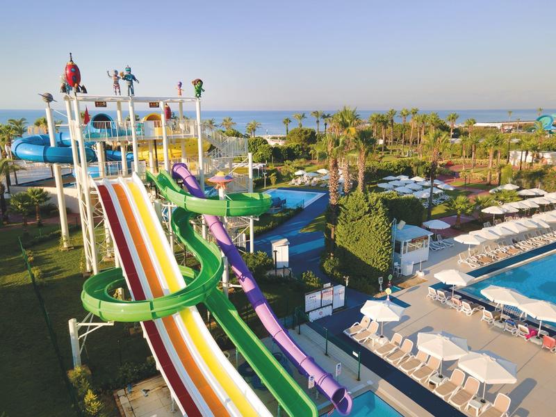 Toboganes de agua coloridos y piscinas en un parque acuático soleado con tumbonas y palmeras.