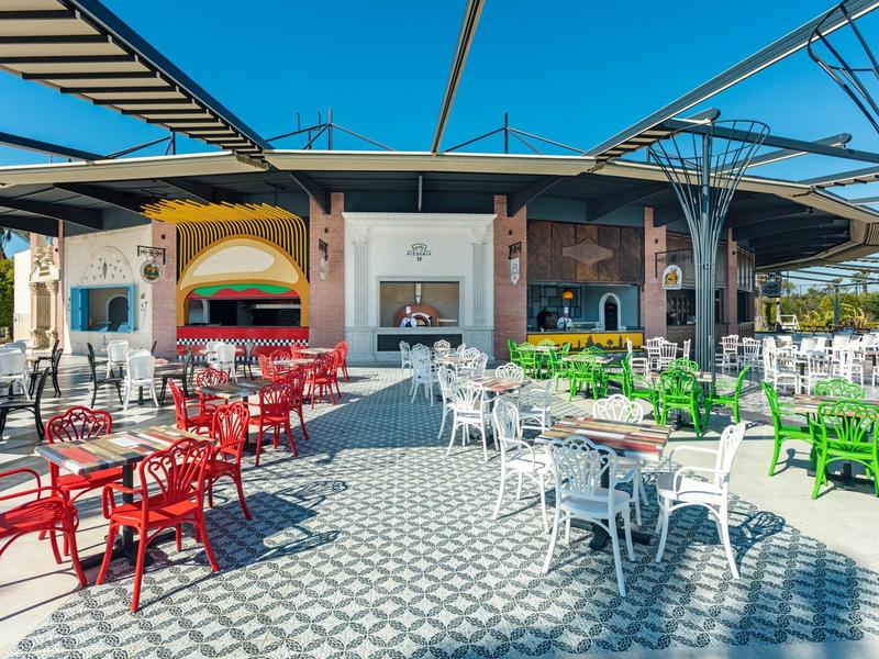Terraza exterior con mesas y sillas en rojo, blanco y verde bajo un techo de estructura metálica.