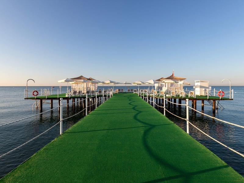 Un muelle largo con alfombra verde conduce a pabellones sobre un mar tranquilo bajo un cielo despejado.