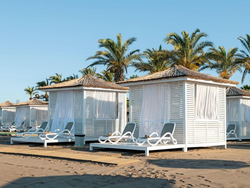 Cabañas blancas en la playa con tumbonas frente a palmeras bajo un cielo despejado.