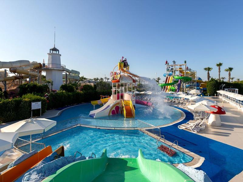 Parque acuático con toboganes y piscina bajo un cielo despejado junto a un faro blanco.