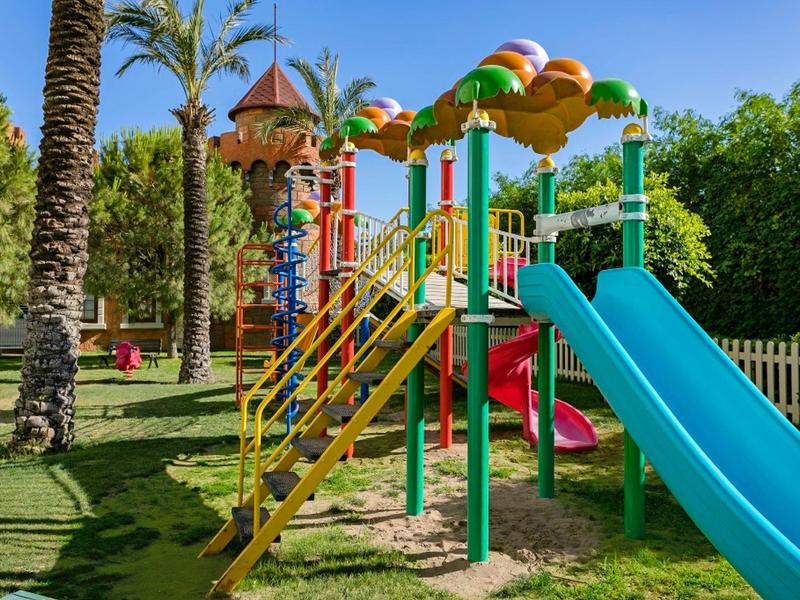 Área de juegos colorida con tobogán azul y estructuras para escalar rodeada de césped y árboles.