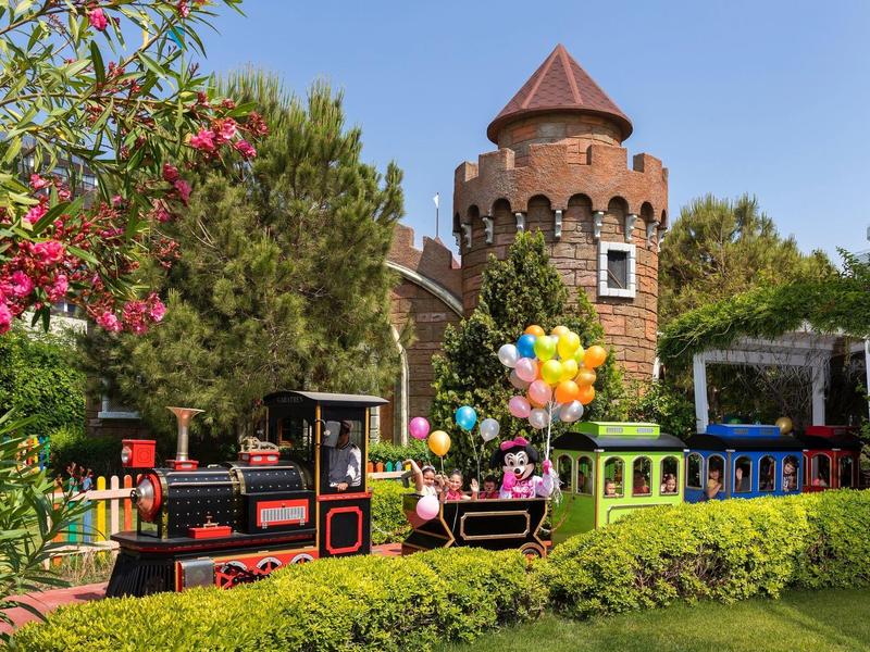 Tren de parque de atracciones colorido frente a un castillo con torre rodeado de árboles y flores.