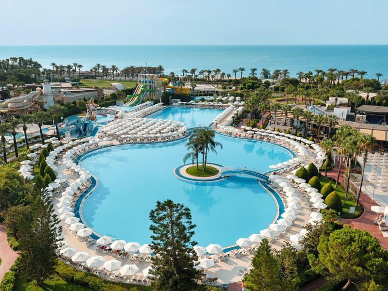 Gran piscina de hotel con muchas tumbonas blancas y palmeras, rodeada de jardines y vista al mar.