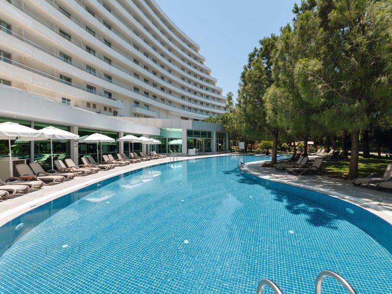Gran piscina de hotel con tumbonas a lo largo y árboles al fondo bajo un cielo soleado.