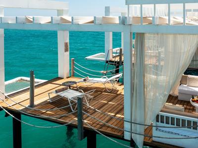 Wooden platform over water with cozy seating and white curtains by the sea.