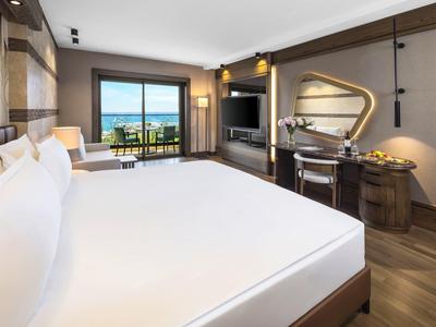 Modern hotel room with large bed, desk, TV, and sea view through the terrace.