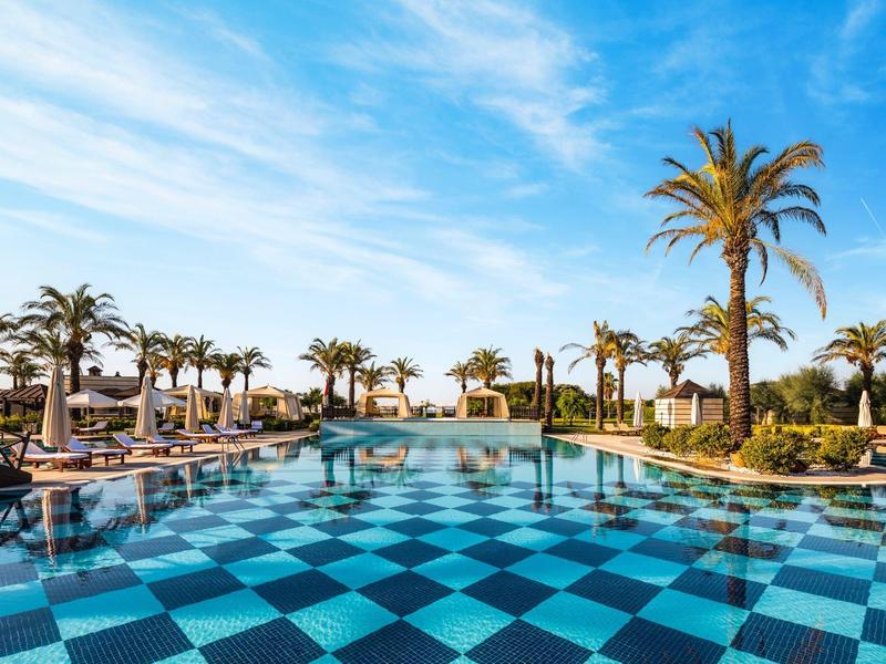 Large pool with blue checkerboard pattern, surrounded by palm trees and loungers under a blue sky.