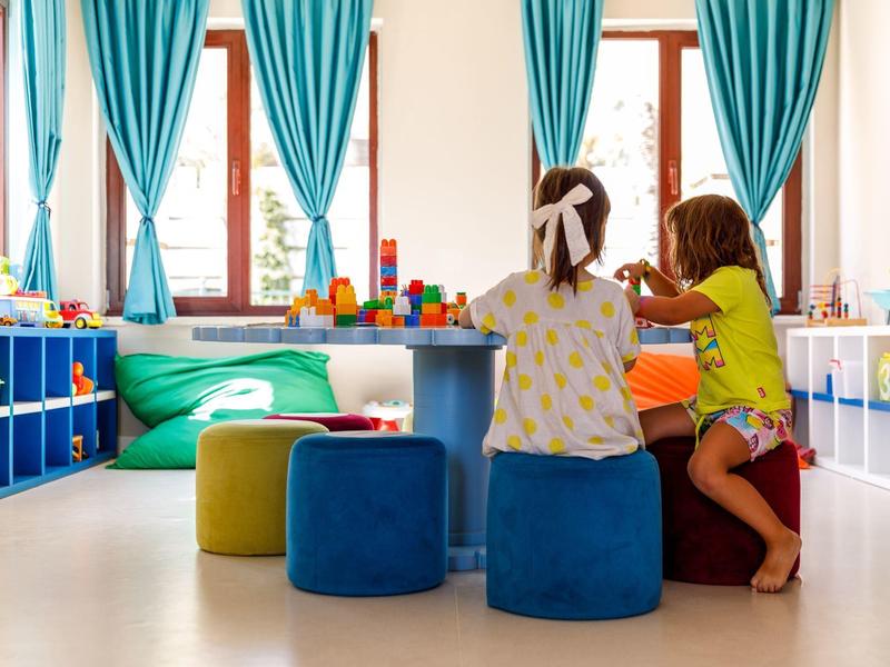 Children play in a bright, colorful playroom with large windows and blue curtains.