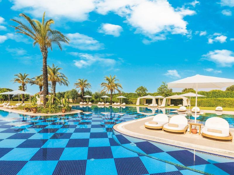 Luxury pool area with blue patterned floor, palm trees, and white sun loungers under blue sky.