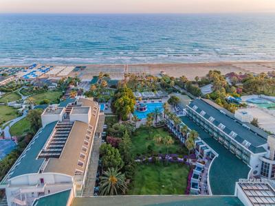 Vue aérienne d'un hôtel en bord de mer avec piscine, jardin et accès direct à la mer au coucher du soleil.