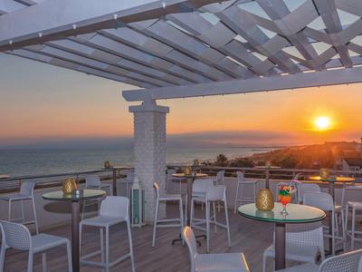 Terrasse avec tables et chaises offrant une vue sur le coucher de soleil sur la mer avec la ville en arrière-plan.