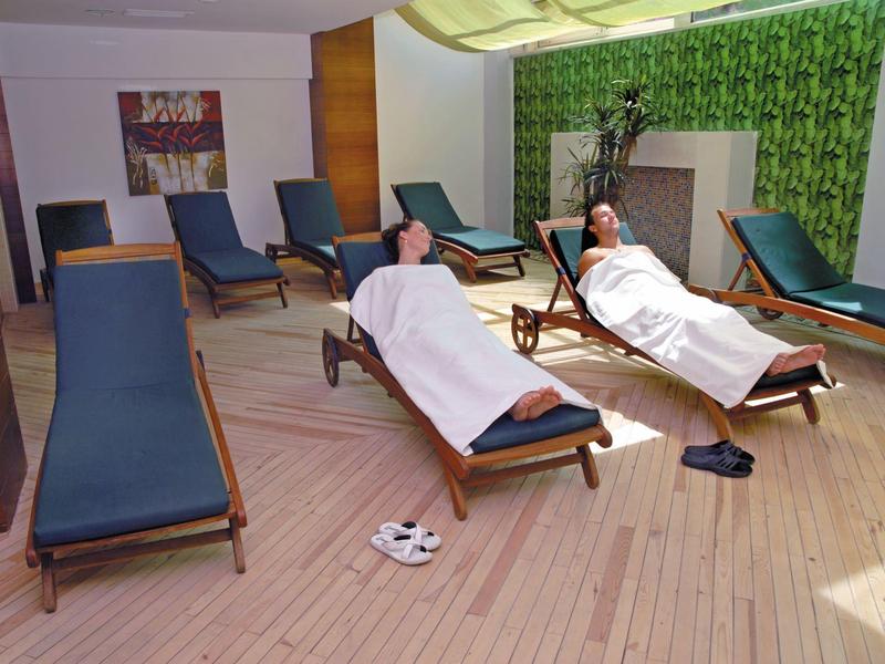 Deux personnes se détendent sur des chaises longues dans une salle de spa lumineuse avec sol en bois et mur à motif vert.