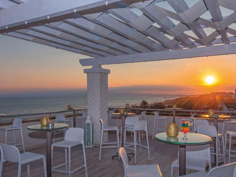 Terrasse avec tables et chaises offrant une vue sur le coucher de soleil sur la mer avec la ville en arrière-plan.
