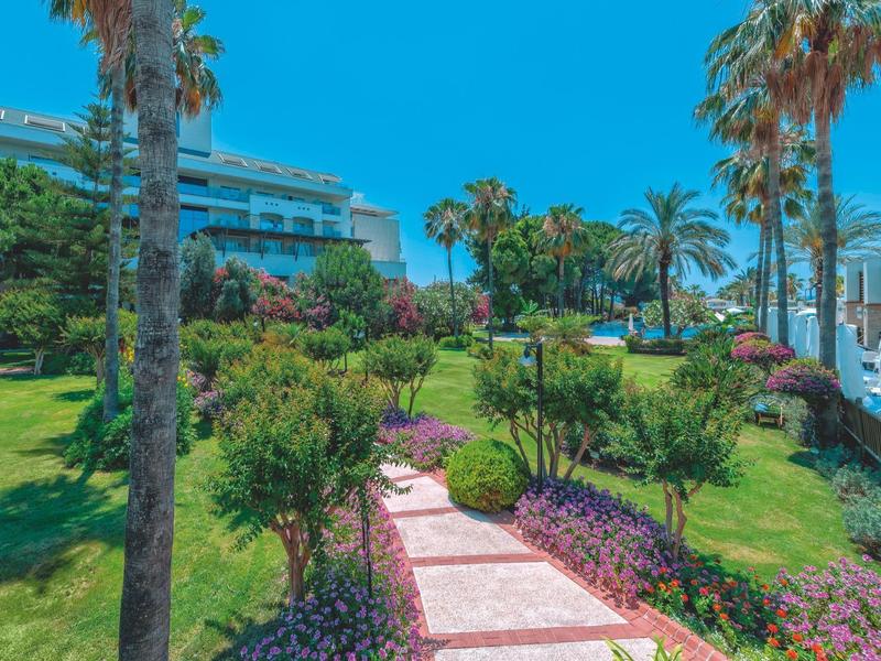 Jardin bien entretenu avec palmiers et fleurs le long d'un chemin menant à un bâtiment hôtelier sous un ciel dégagé.