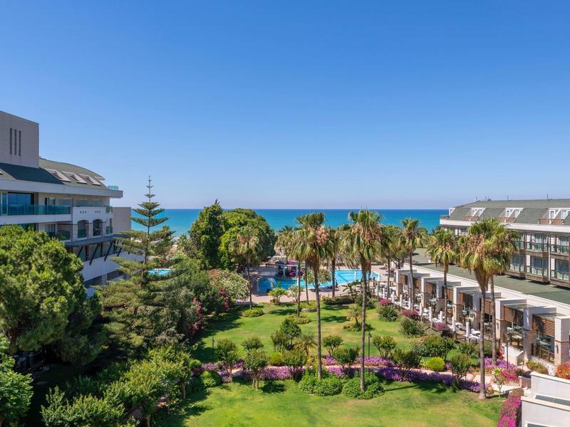 Vue sur un hôtel avec jardin, palmiers, piscine et mer sous un ciel bleu clair.