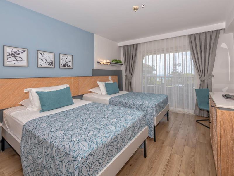 Chambre d'hôtel lumineuse avec deux lits simples, bureau et grandes fenêtres avec rideaux.