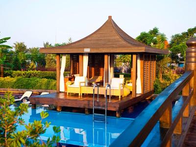 Houten paviljoen met zitplaatsen bij een blauw zwembad omgeven door tropische planten en een houten brug.
