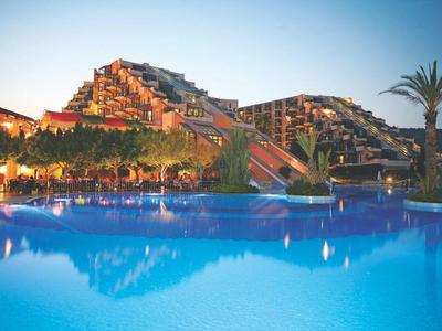 Hotel moderno con edifici a terrazze su più piani accanto a una piscina blu al tramonto.
