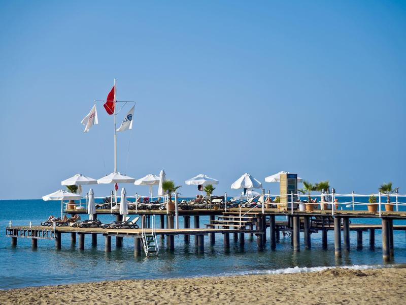 Houten pier met witte parasols en vlaggen die uitsteekt over kalme zee onder een heldere blauwe lucht.