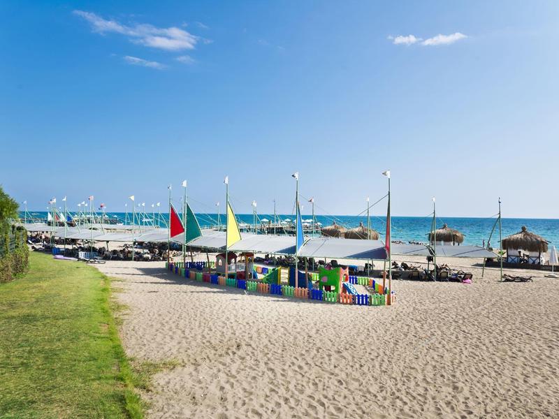 Zonnig strand met kleurrijke parasols, ligbedden en vlaggen bij rustige zee en heldere lucht.