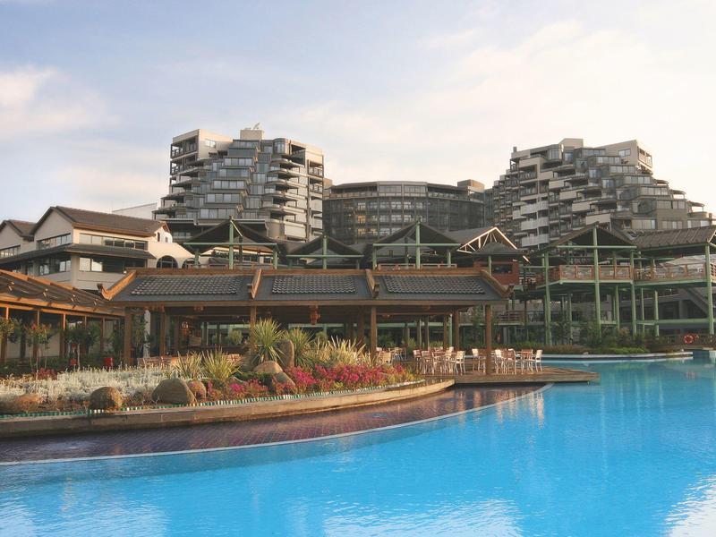 Moderne hotellgebouwen met groot zwembad en aangelegd tuingebied ervoor.