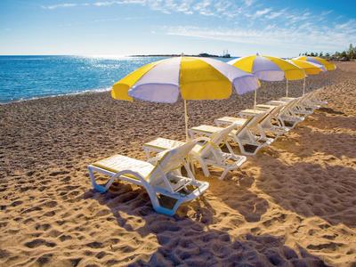Reihe gelb-weißer Sonnenschirme mit Liegestühlen auf Sandstrand am blauen Meer unter klarem Himmel.