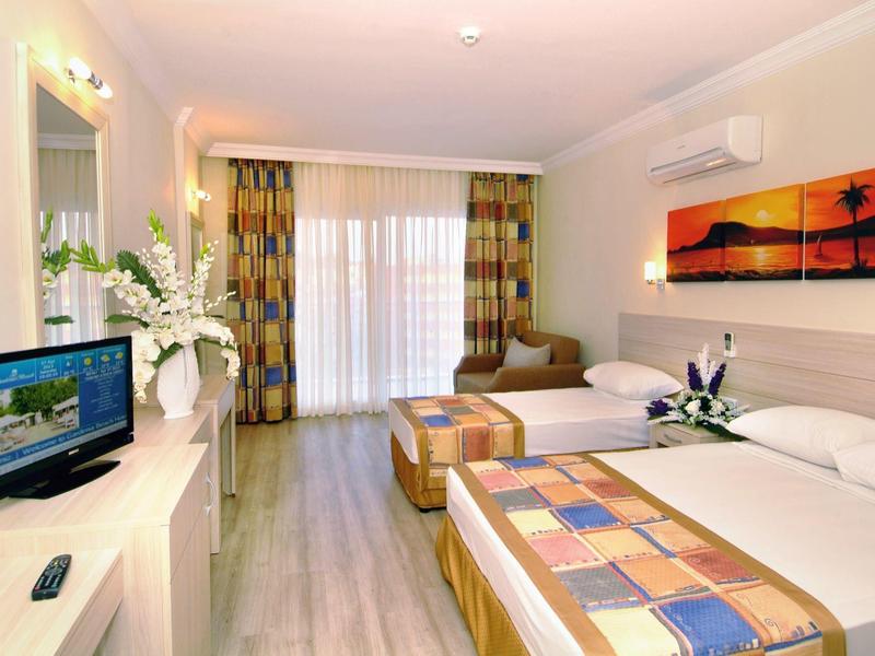 Helles Hotelzimmer mit zwei Betten, Fernseher, Blumen und großem Fenster mit Vorhängen.