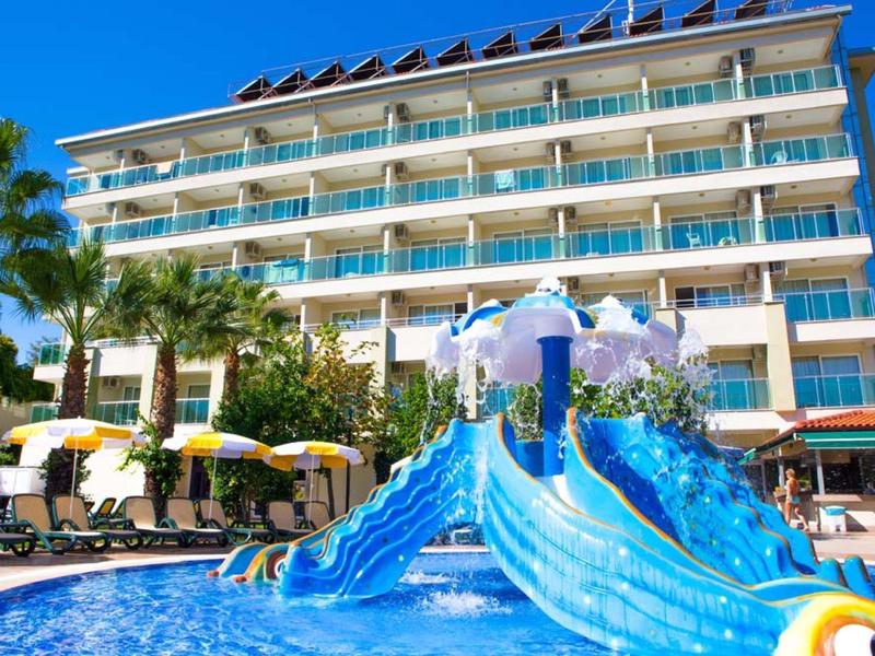 Hotel mit Wasserrutsche vor blauem Himmel und Palmen, Liegestühle und Sonnenschirme am Pool.
