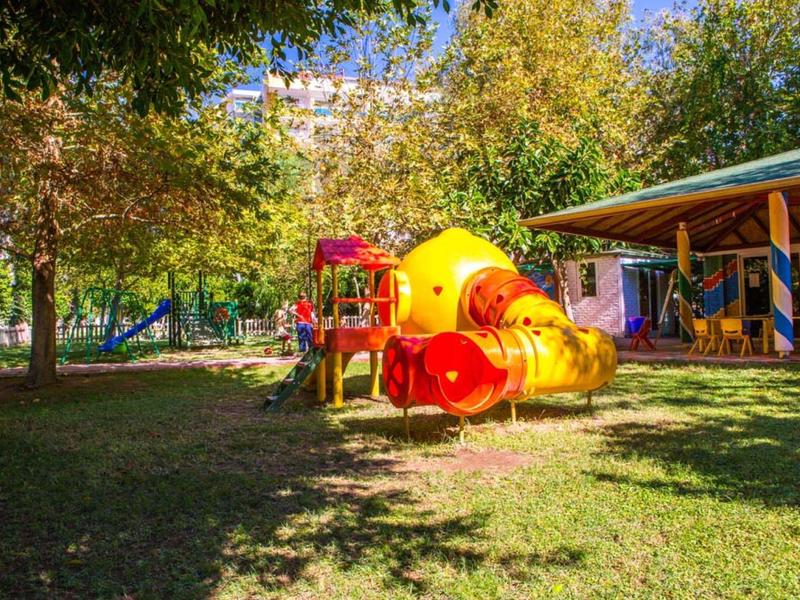 Ein bunter Spielplatz mit Rutsche und Tunnelröhre, umgeben von grasbewachsenem Boden und Bäumen.