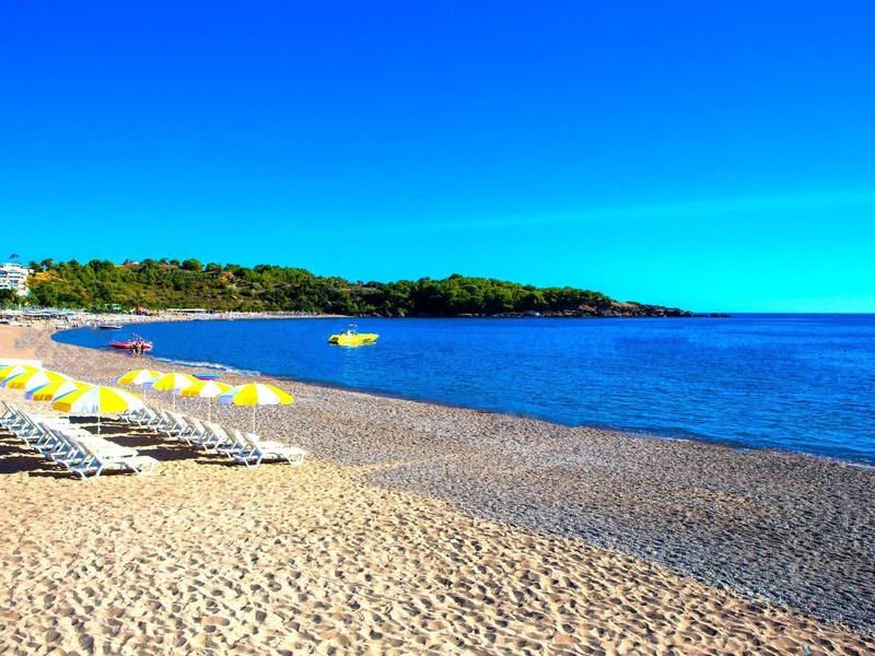 Sonniger Strand mit Sand, leeren Liegestühlen und klarem, blauem Meer unter blauem Himmel.