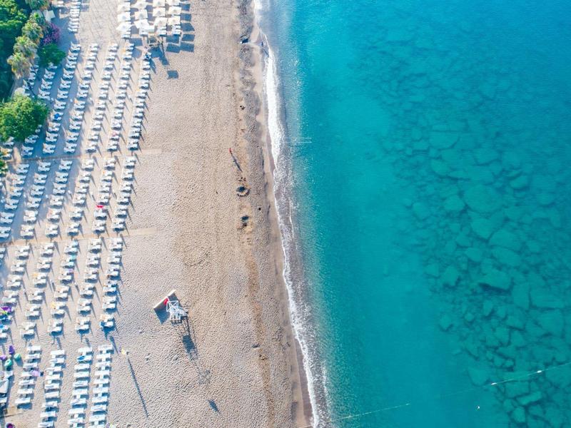 Luftaufnahme eines Sandstrands mit Reihen von Sonnenschirmen und Liegestühlen neben klarem, blauem Meer.