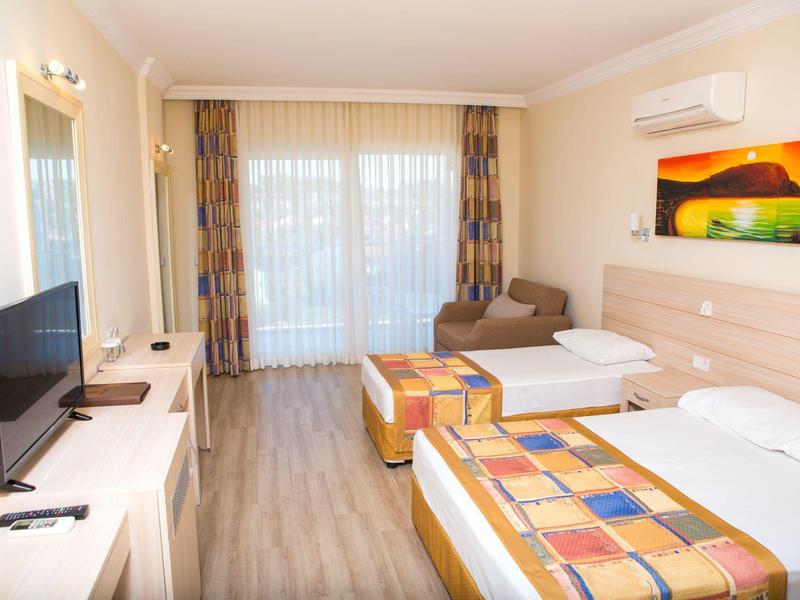 Helles Hotelzimmer mit zwei Betten, Fernseher, Sofa, Klimaanlage und großen Fenstern mit Vorhängen.