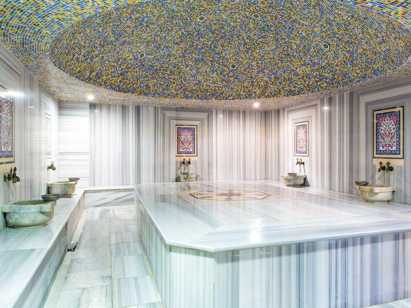 Heller, großer Hamam mit Marmorwänden, zentralem Becken und farbenfroher Deckenmosaik.