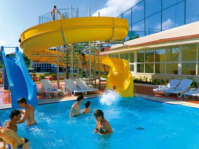 Schwimmbecken mit gelber und blauer Wasserrutsche, spielende Kinder, Sonnenterrasse mit Liegestühlen.