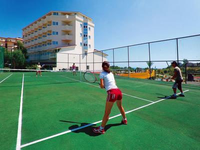 Zwei Personen spielen Tennis auf einem grünen Platz bei klarem Himmel und Gebäuden im Hintergrund.