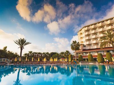 Hotel mit Palmen und Sonnenliegen neben großem Pool unter blauem Himmel mit Wolken.