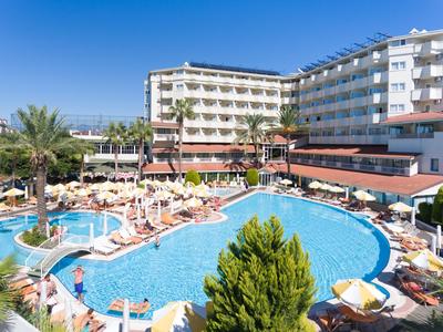 Hotel s velkým venkovním bazénem, lehátky kolem a palmami pod jasně modrou oblohou.