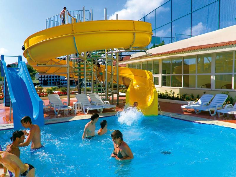 Children playing in a pool with yellow and blue water slides beside a building with glass façade.