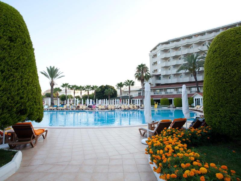 View of a hotel pool with lounge chairs, palm trees, and a multi-story building in the background.