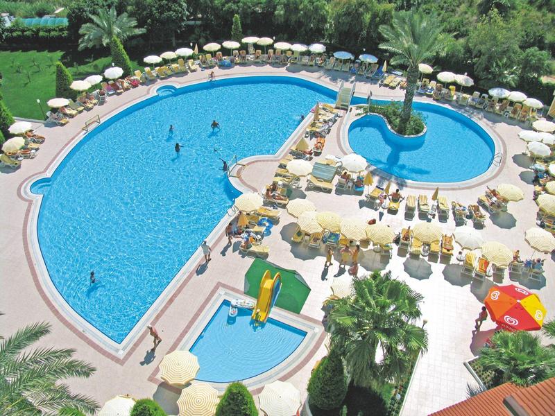 Large hotel pool with sun loungers and shaded areas, surrounded by palm trees and greenery.