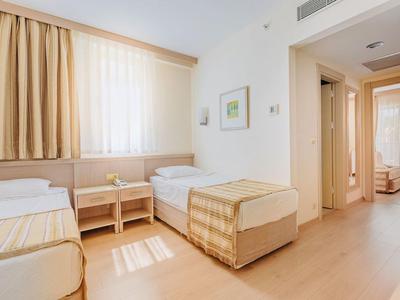 Bright hotel room with two single beds, wooden floor, and beige curtains.