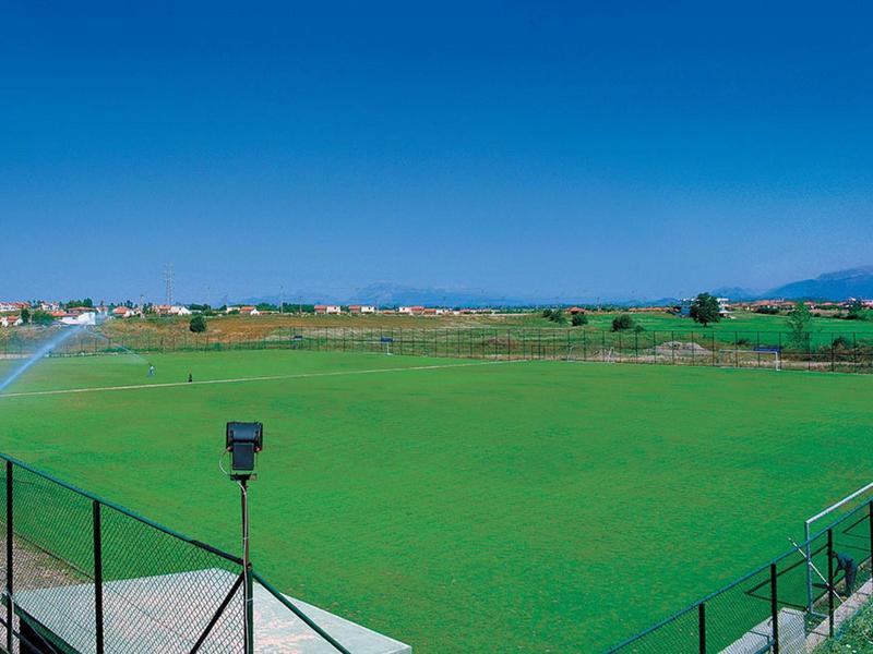 Wide green sports field under clear blue sky with sprinklers and distant buildings.