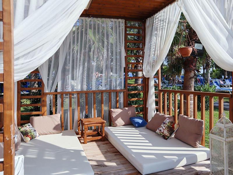 Cozy wooden outdoor lounge with white drapes, cushions, and a small rocking chair under a shaded patio.
