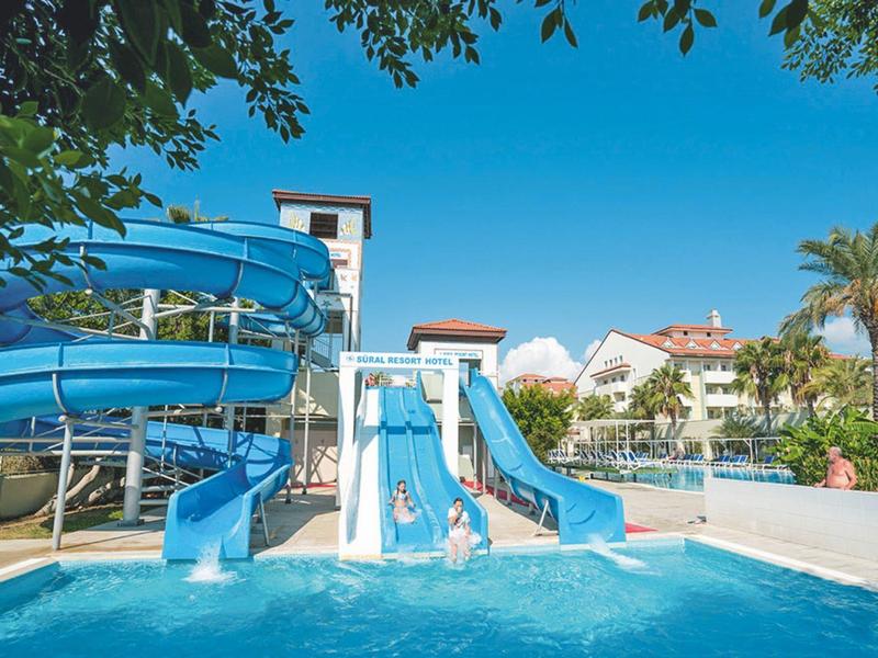 Water slides leading into a clear blue pool at a sunny resort with palm trees and buildings.