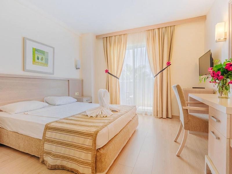 Bright hotel room with double bed, desk, TV, and large window with beige curtains.
